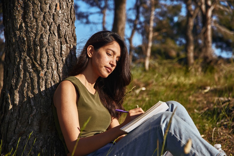 A student leans against a tree while writing in a journal.