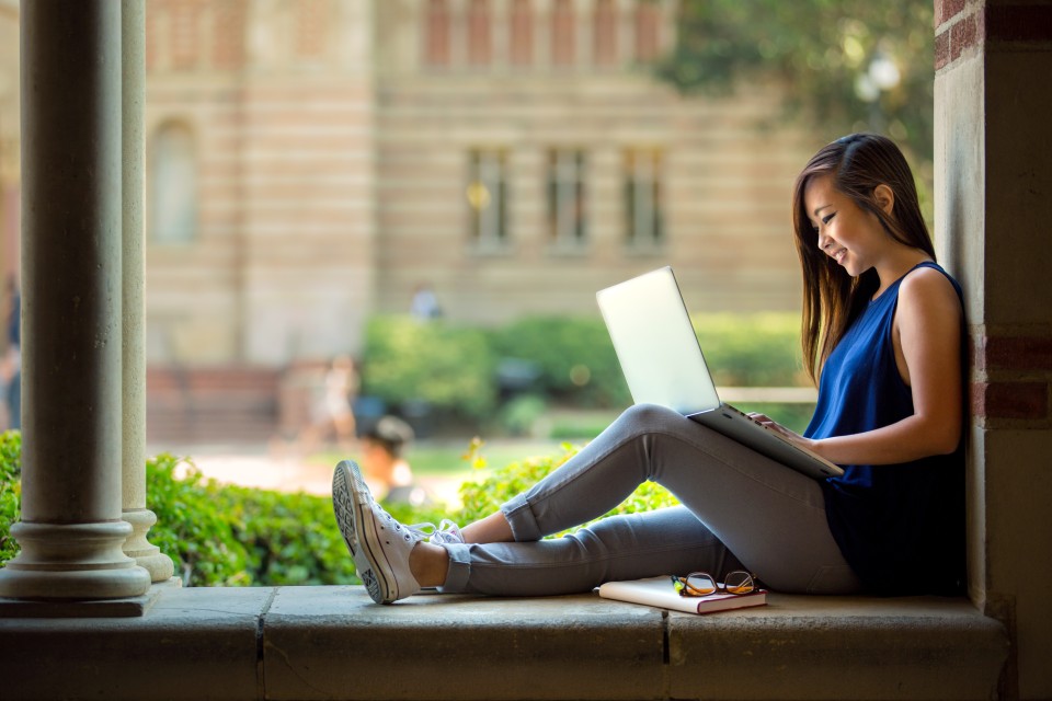 A college student works on a laptop outside a campus building.