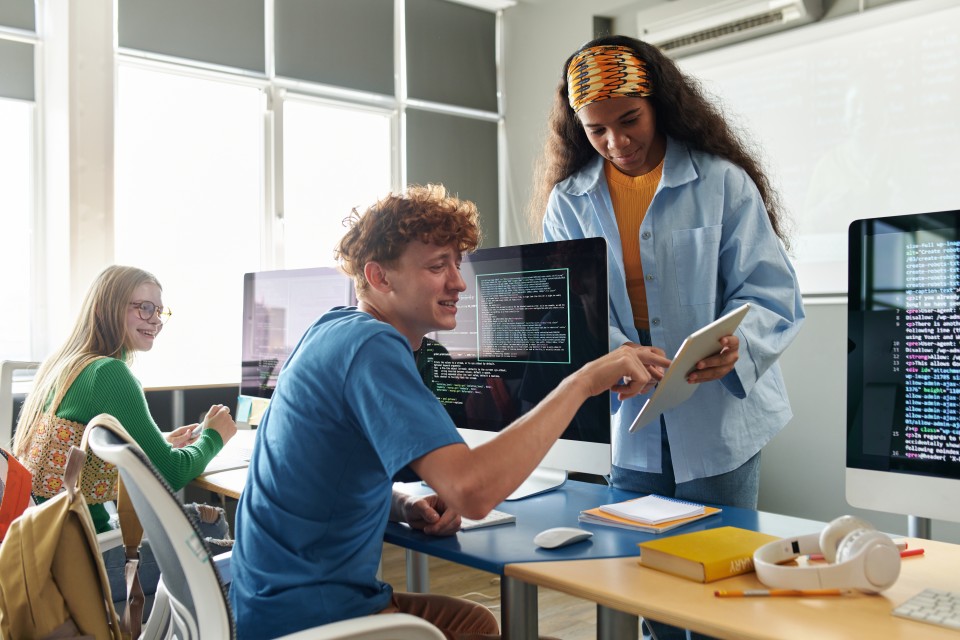 Students working in front of large computer monitors. The man is pointing at something on another student's tablet computer.