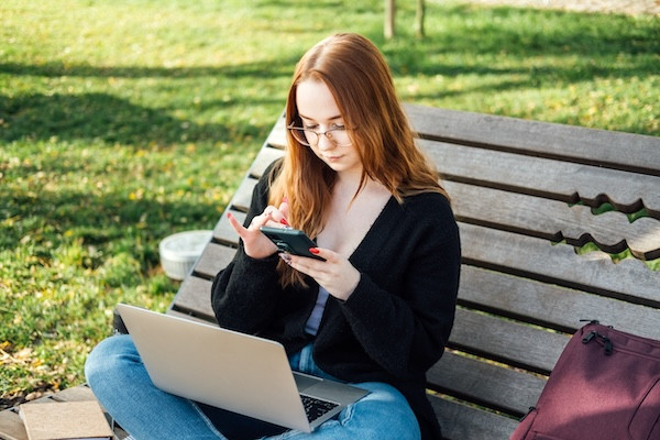 A student sits outdoors on a bench, typing on a phone with a laptop in her lap.
