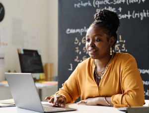 Instructor in front of blackboard looking at a laptop.